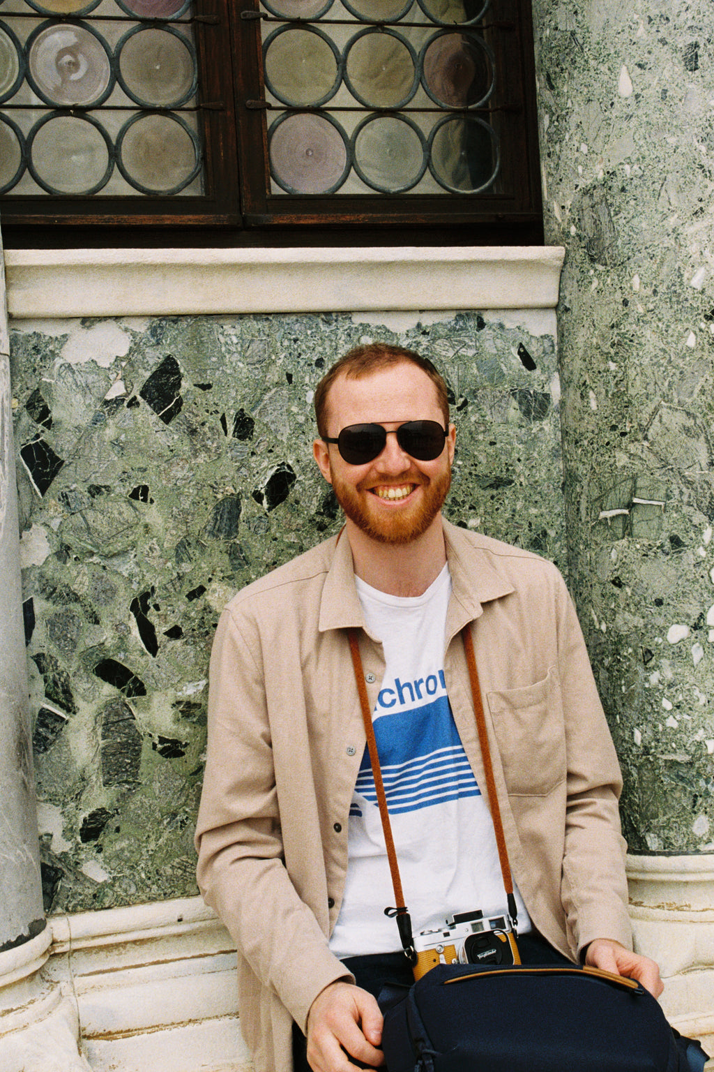 Man wearing sunglasses and a beige coat standing on a stone floor.