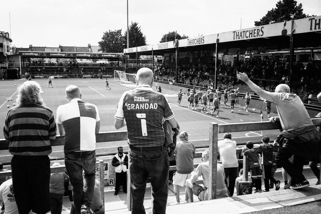 Soccer match with fans in the stands, one fan wearing a 'Grandad' jersey.
