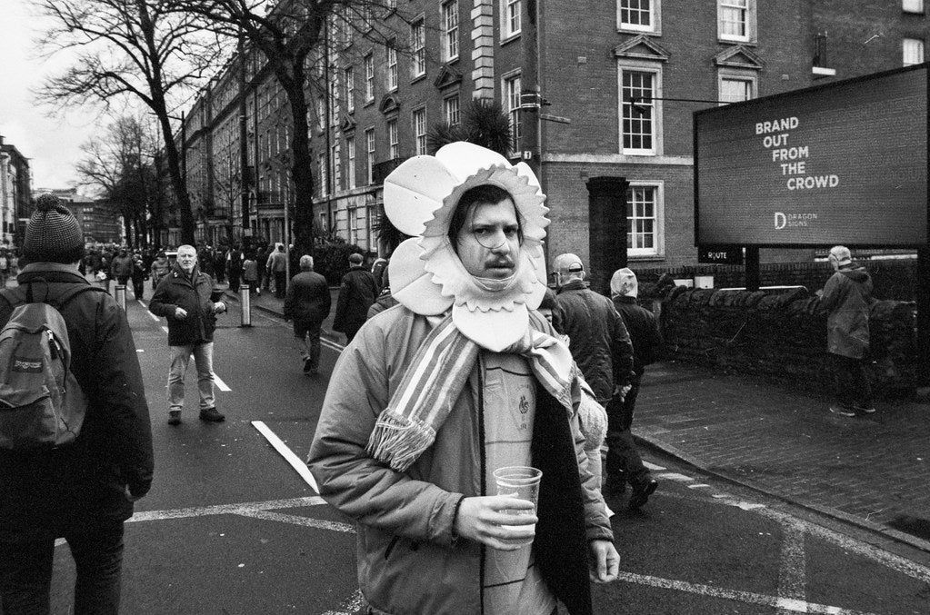 Person in vintage costume holding a drink on a city street with pedestrians and a digital display in the background.